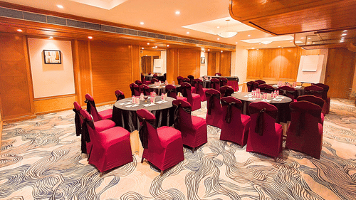 A well-lit banquet hall at Pride Plaza Ahmedabad, featuring round tables with red tablecloths, chairs with red covers, and a marble floor.